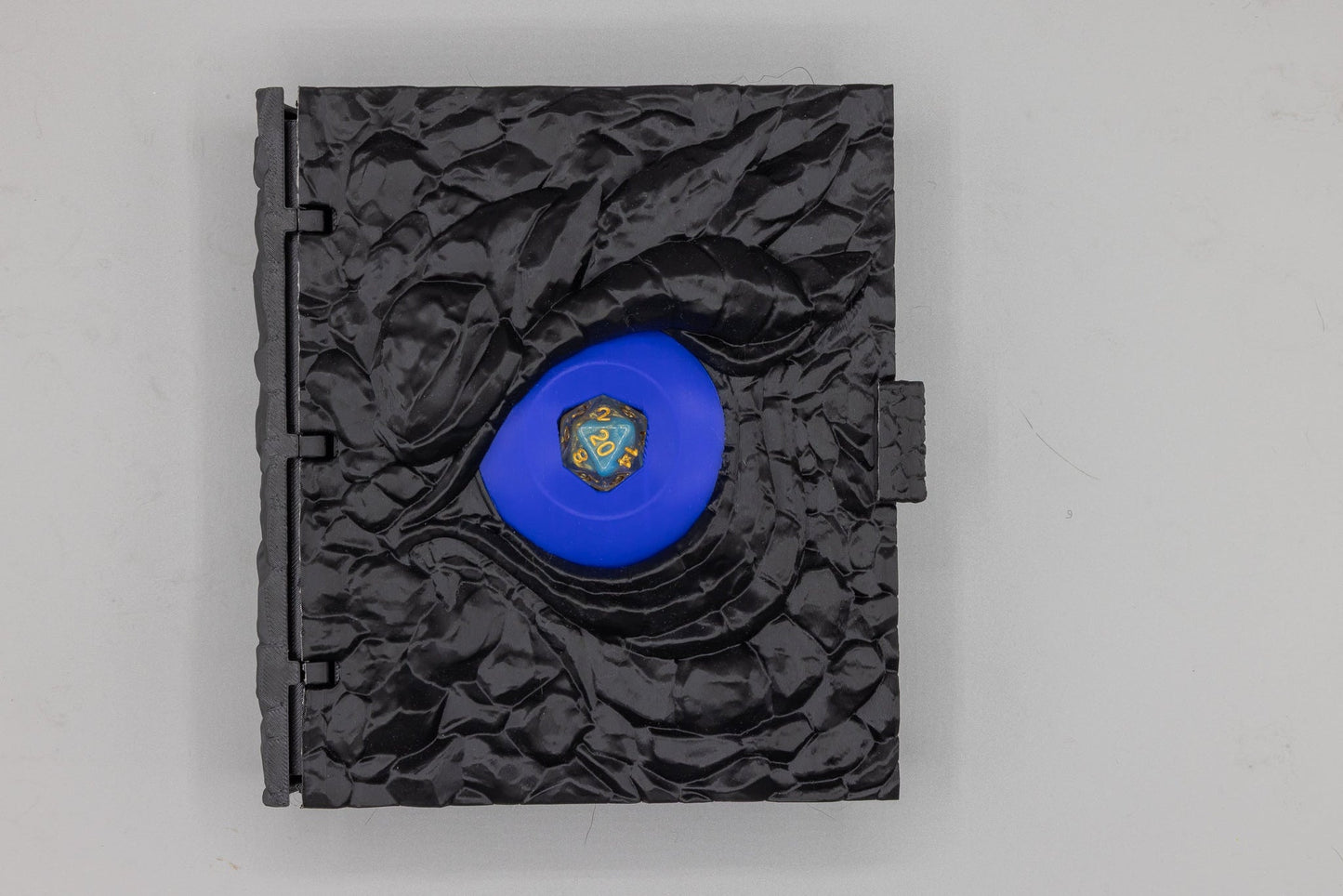 Dragon Dice Book closed — black dragon-scale cover with an electric-blue dragon eye holding a d20