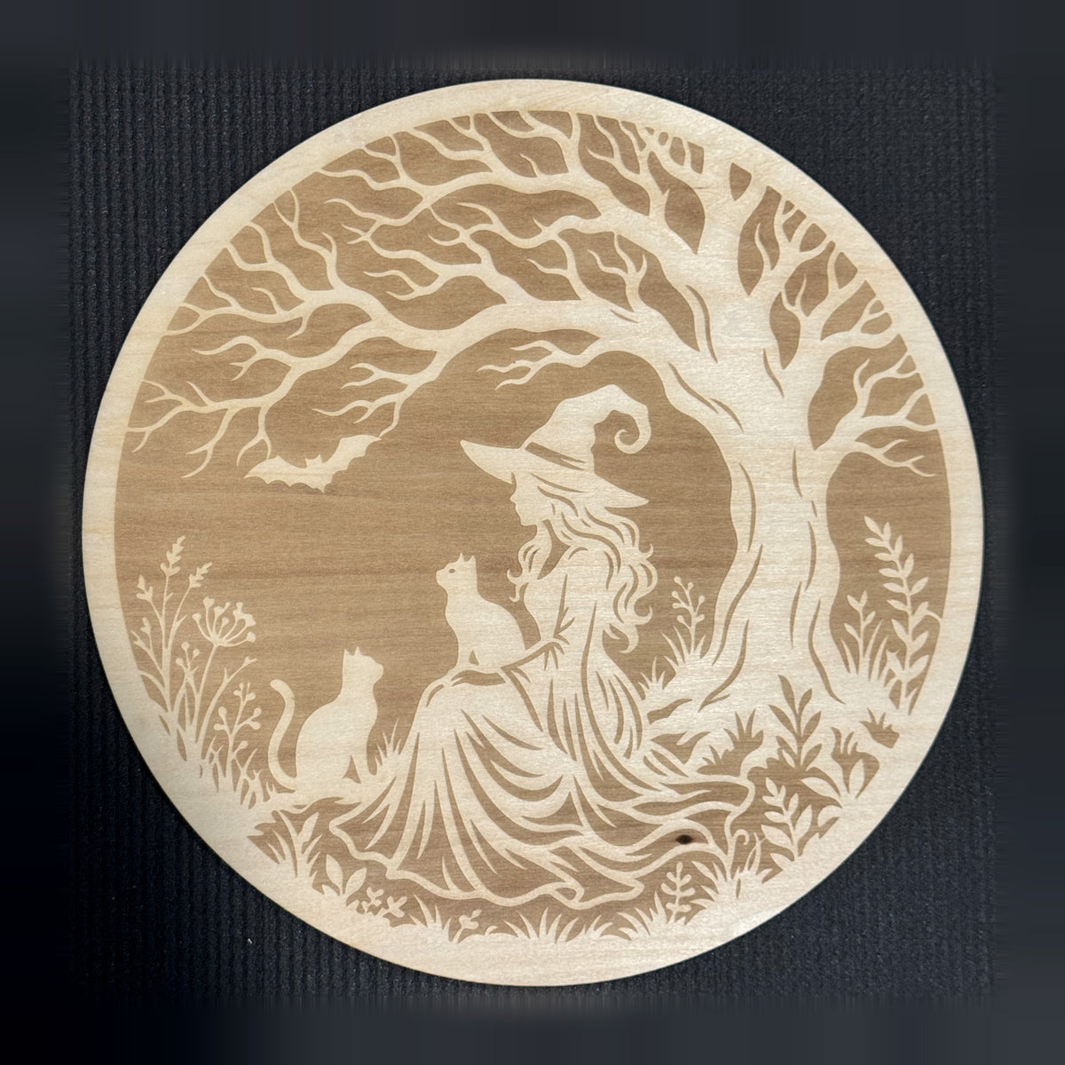 Under the Witching Tree — 11 inch round natural basswood wall art engraved with a witch seated beneath a twisted tree, two cats and a bat, framed by wildflowers